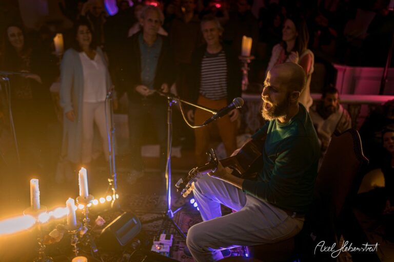 Musiker mit Gitarre spielt in einer stimmungsvollen, beleuchteten Umgebung vor einem begeisterten Publikum – © Axel Hebenstreit