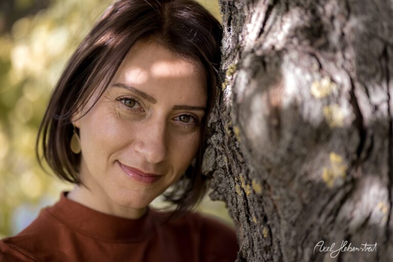 Junge Frau lehnt lächelnd an einem Baum und genießt die warmen Sonnenstrahlen im Freien – © Axel Hebenstreit