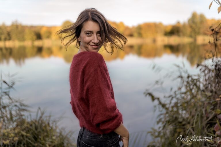 Lächelnde Frau mit wehendem Haar am Ufer eines ruhigen Sees in herbstlicher Landschaft – © Axel Hebenstreit
