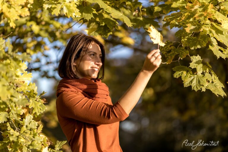 Junge Frau in herbstlicher Landschaft hält ein Blatt in der Hand und lächelt, umgeben von bunten Blättern – © Axel Hebenstreit