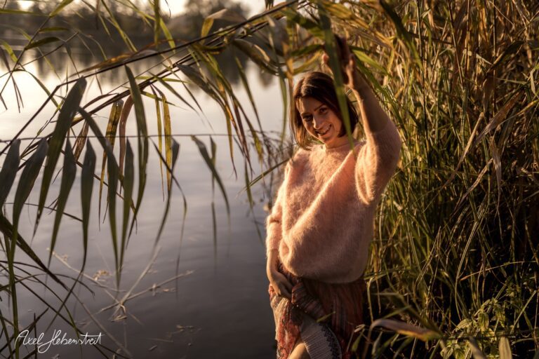 Eine Braunhaarige Frau in einem weichen Pullover posiert lächelnd zwischen Schilfpflanzen am Wasser – © Axel Hebenstreit