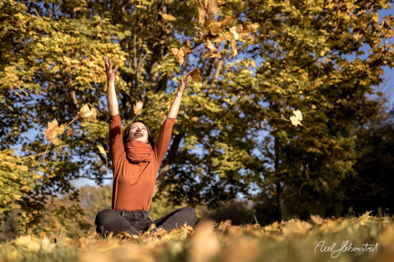 Frau sitzt im bunten Herbstlaub und wirft mit Freude Blätter in die Luft – © Axel Hebenstreit