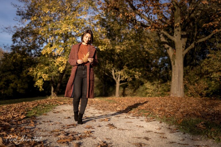 Eine Frau in einem herbstlichen Park geht auf einem mit Laub bedeckten Weg und hält ein Blatt in der Hand – © Axel Hebenstreit