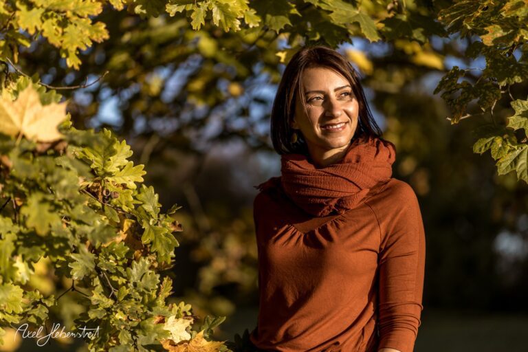 Eine lächelnde Frau in einem herbstlichen Umfeld mit bunten Blättern im Hintergrund – © Axel Hebenstreit.