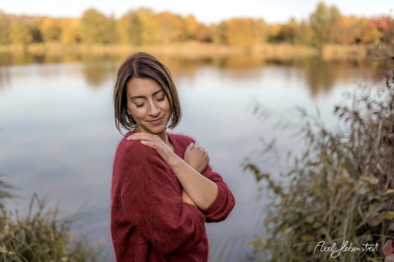 Frau in weichem Pullover steht am Ufer eines ruhigen Sees und genießt die herbstliche Natur – © Axel Hebenstreit