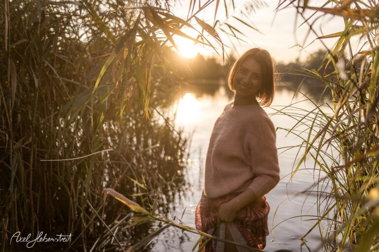 Junge Frau mit lächelndem Gesicht steht am Ufer eines Sees, umgeben von Schilf und Sonnenlicht – © Axel Hebenstreit