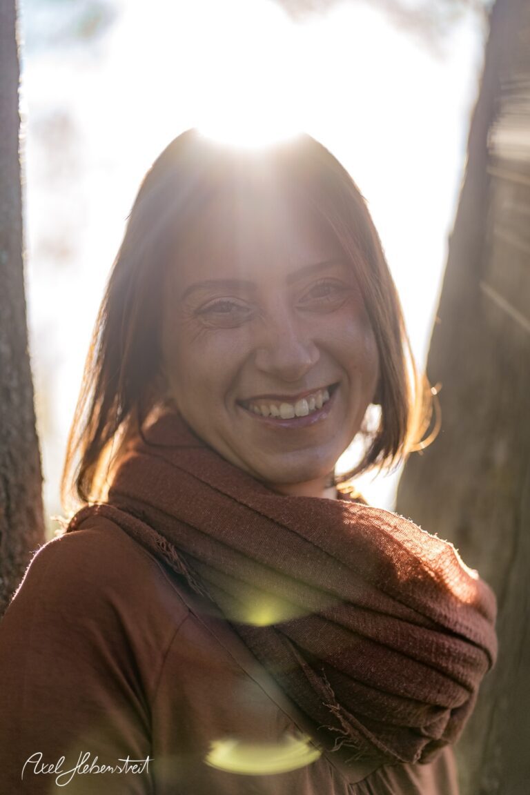 Lächelnde Frau mit Sonnenstrahlen im Hintergrund, umgeben von Bäumen, die eine freundliche und entspannte Stimmung vermittelt – © Axel Hebenstreit