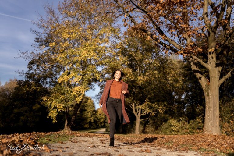 Eine Frau in Herbstkleidung läuft fröhlich auf einem bunten, von Bäumen gesäumten Weg entlang. – © Axel Hebenstreit