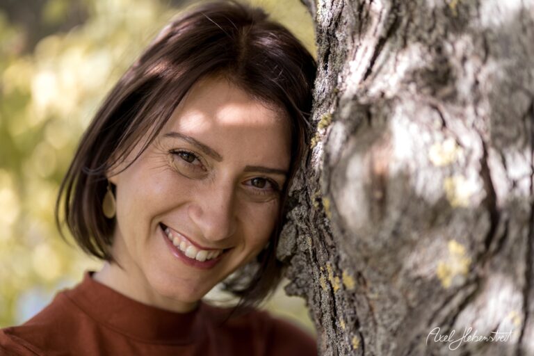 Frau lächelt verträumt neben einem Baum in der Natur, umgeben von warmem Licht – © Axel Hebenstreit