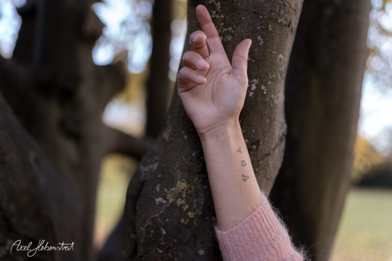 Eine Hand mit zarten Tattoos berührt einen Baumstamm in einer herbstlichen Landschaft. – © Axel Hebenstreit