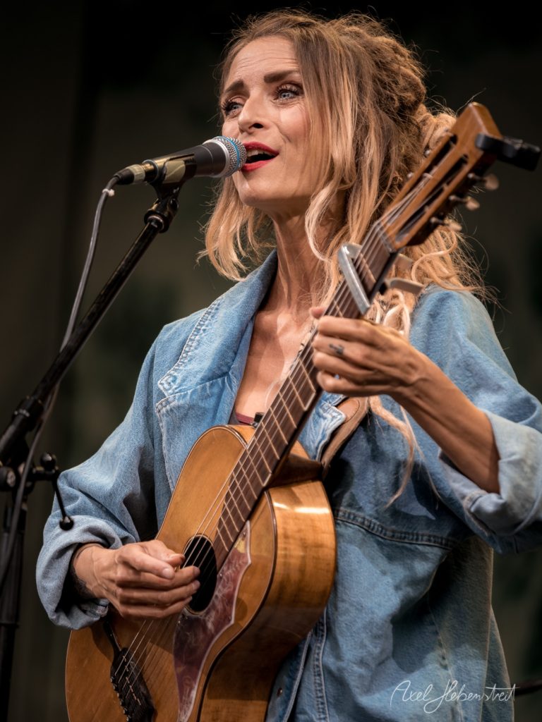 Eine Sängerin mit Gitarre in einem lässigen Denim-Jacket singt leidenschaftlich auf einer Bühne – © Axel Hebenstreit