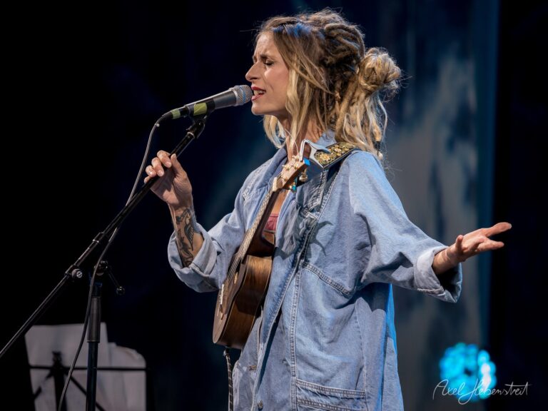 Eine Sängerin mit Gitarre und Dreadlocks performt leidenschaftlich auf einer Bühne. – © Axel Hebenstreit