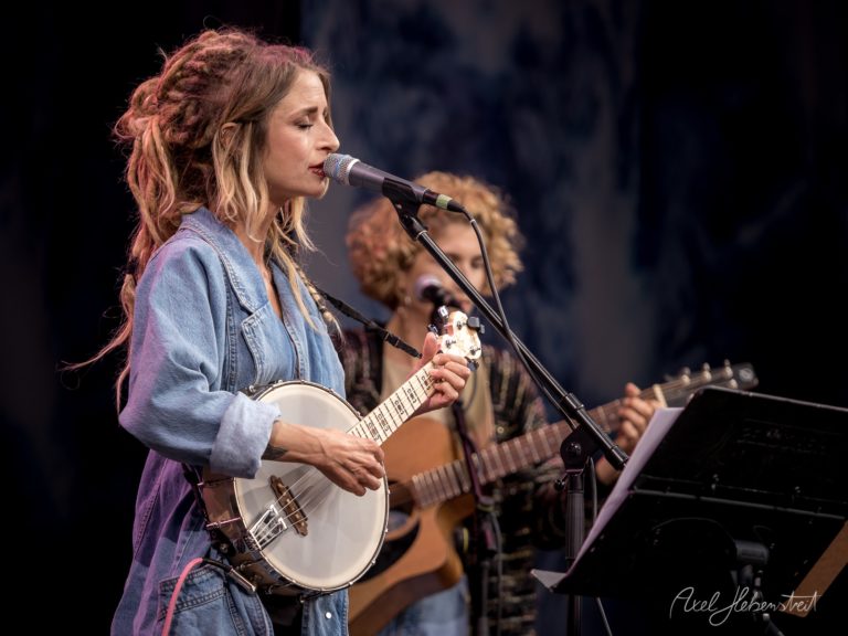 Frau mit Halskrause spielt Banjo und singt, während ein Musiker im Hintergrund mit Gitarre begleitet – © Axel Hebenstreit