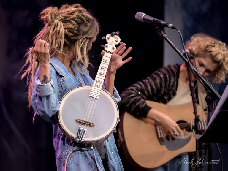 Frau mit Dreadlocks spielt Banjo und zeigt eine lebendige Performance auf der Bühne – © Axel Hebenstreit