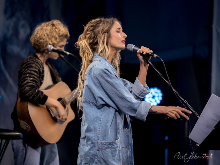 Zwei Sängerinnen performen leidenschaftlich auf einer Bühne, während das Publikum gespannt lauscht – © Axel Hebenstreit