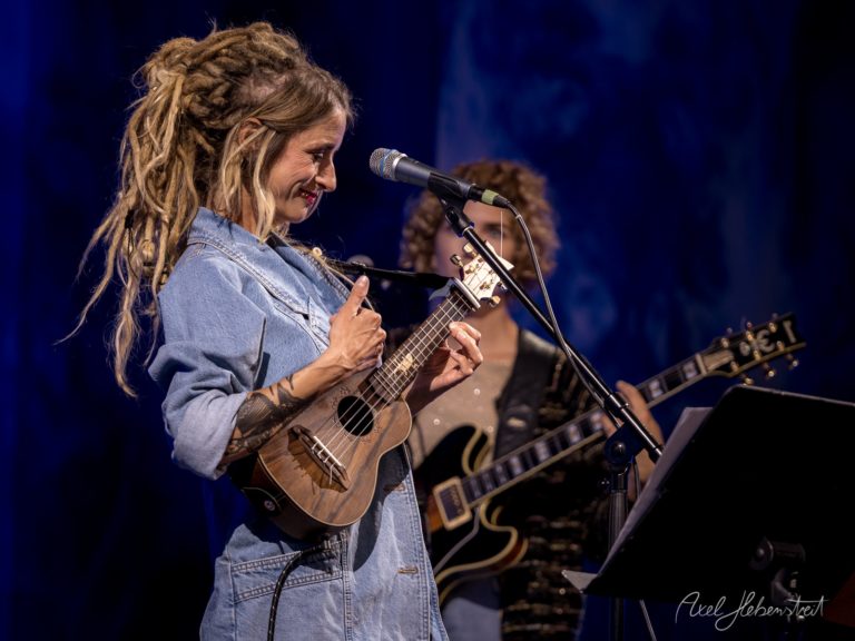 Frau mit Dreadlocks spielt Ukulele auf der Bühne und lächelt begeistert ins Publikum – © Axel Hebenstreit