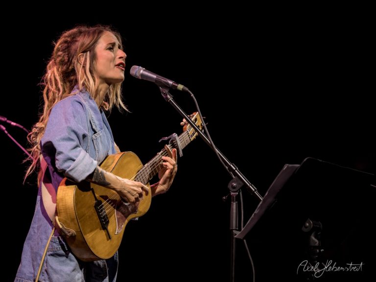 Eine Musikerin mit Gitarre singt leidenschaftlich auf einer Bühne, umgeben von sanften Lichtern – © Axel Hebenstreit