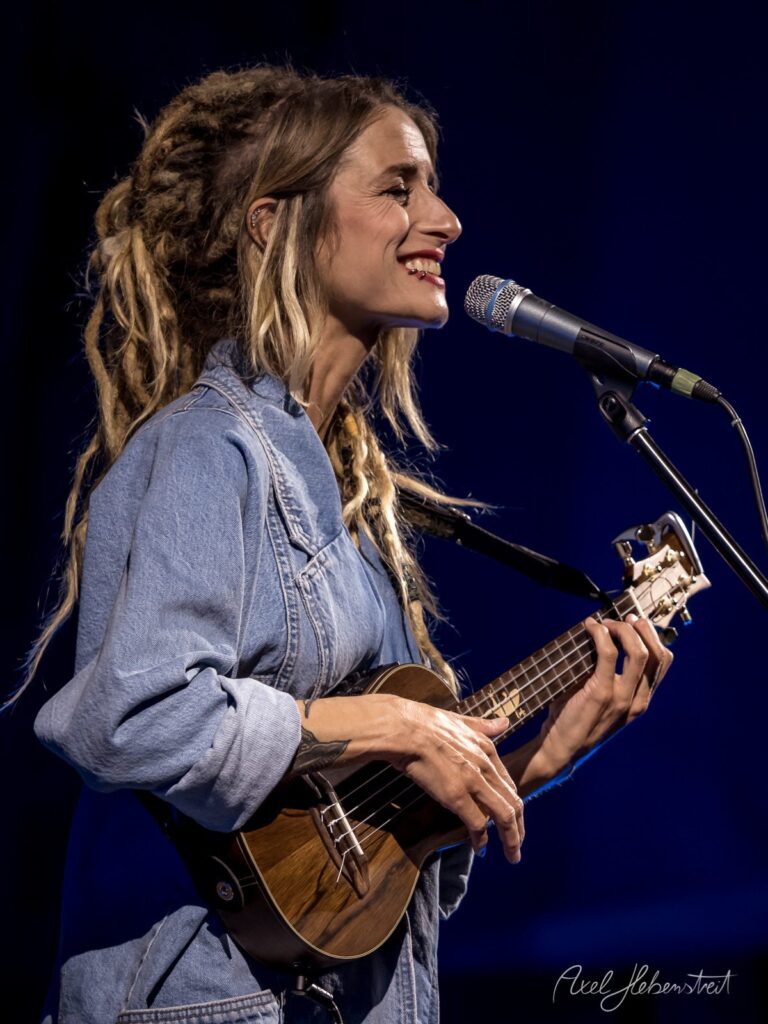 Eine Musikerin mit Dreadlocks spielt begeistert Ukulele und singt in einem atmosphärischen Konzertsaal – © Axel Hebenstreit