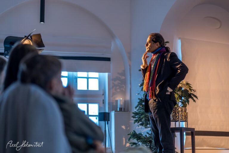 Ein nachdenklicher Redner steht auf einer Bühne, umgeben von natürlichem Licht und festlicher Dekoration – © Axel Hebenstreit