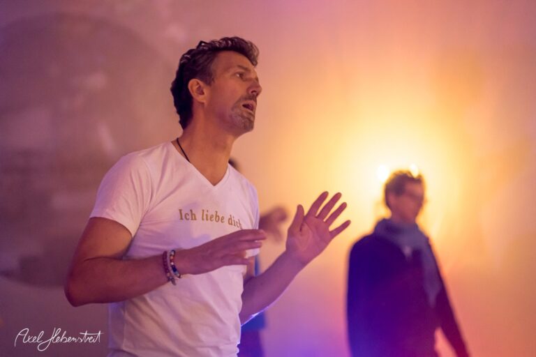 Ein Mann mit leidenschaftlicher Gestik spricht während einer spirituellen Veranstaltung in sanftem Licht – © Axel Hebenstreit