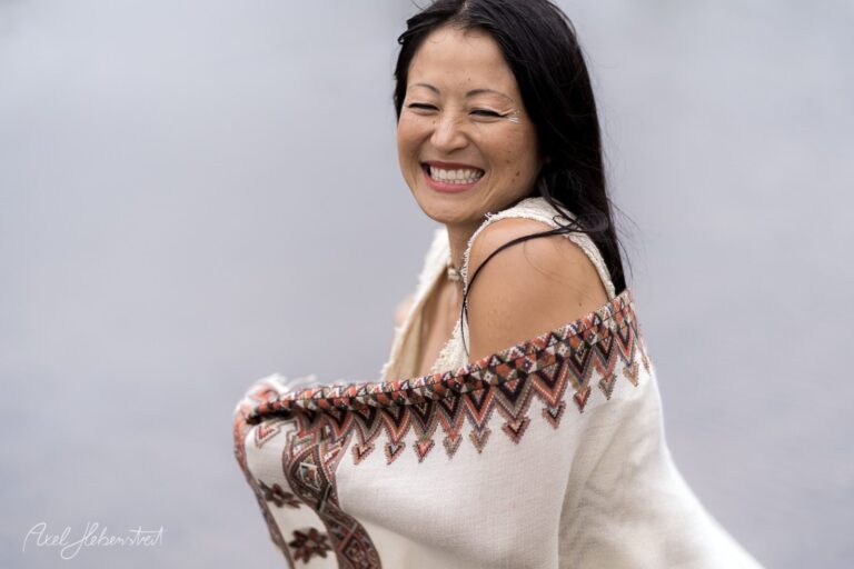 Lächelnde Frau in traditionellem Strickpullover mit Muster blickt über die Schulter vor neutralem Hintergrund – © Axel Hebenstreit