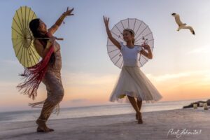 Zwei Frauen tanzen mit Sonnenregenschirmen am Strand bei Sonnenuntergang und versprühen Lebensfreude – © Axel Hebenstreit