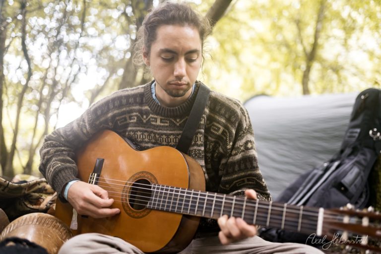 junger Mann spielt an einem ruhigen Ort in der Natur Gitarre, umgeben von Bäumen und Campingausrüstung – © Axel Hebenstreit