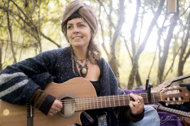 Frau mit Gitarre in natürlicher Umgebung, lächelnd und entspannt, umgeben von Bäumen und sanftem Licht – © Axel Hebenstreit