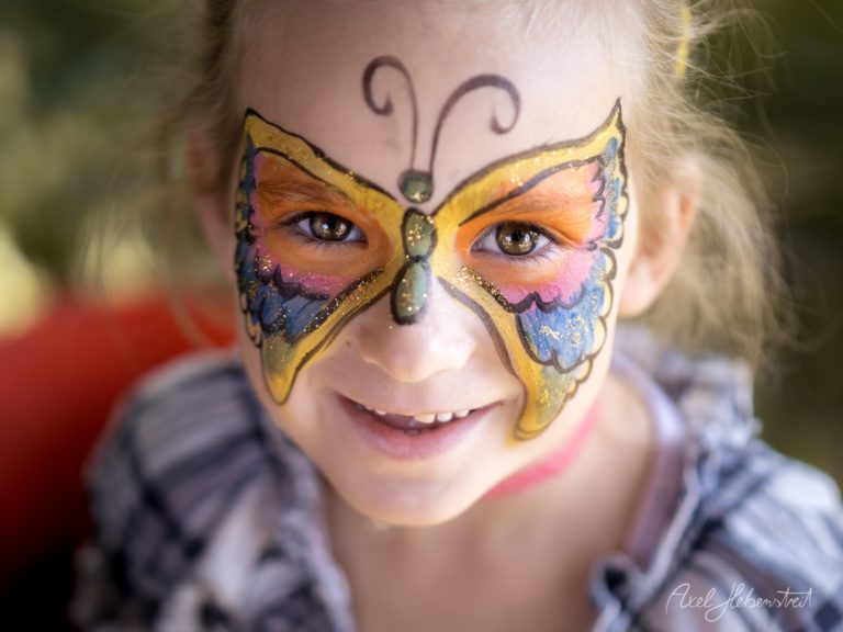 Lächelndes Kind mit aufwendigem Schmetterlings-Gesichtsbemalung in Gold, Orange und Blau mit glitzernden Details – © Axel Hebenstreit