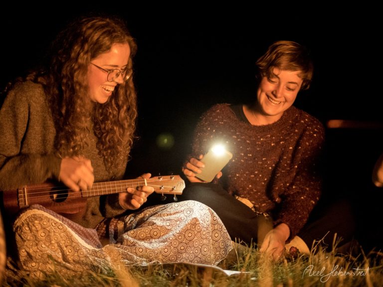 Zwei junge Frauen musizieren nachts am Lagerfeuer, eine spielt Ukulele, die andere hält eine Taschenlampe – © Axel Hebenstreit