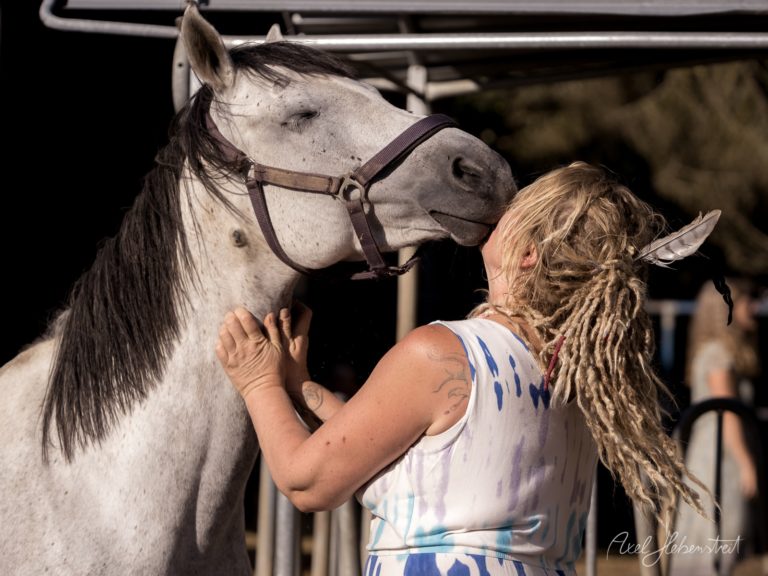 Eine Frau mit Dreadlocks und Flügelkostüm streichelt zärtlich ein weißes Pferd in einem Stall – © Axel Hebenstreit