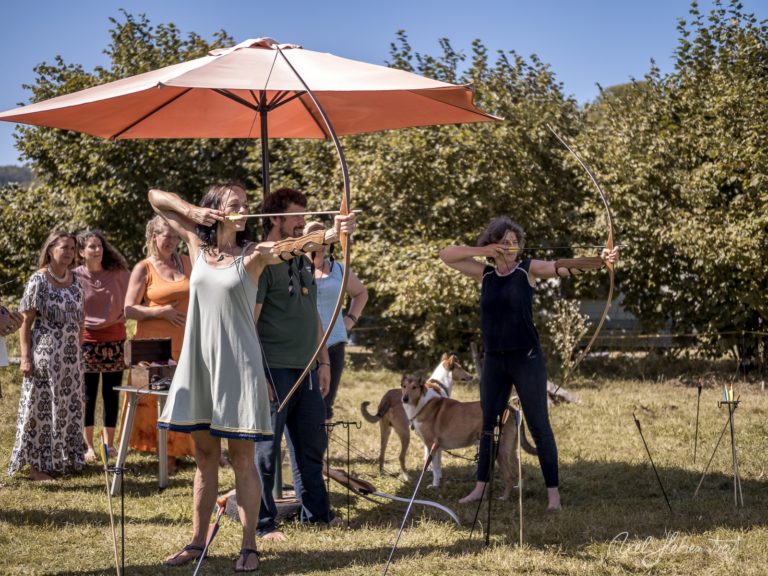 Bogenschießen-Workshop im Freien mit Teilnehmern unter rotem Sonnenschirm in einer grünen Landschaft – © Axel Hebenstreit