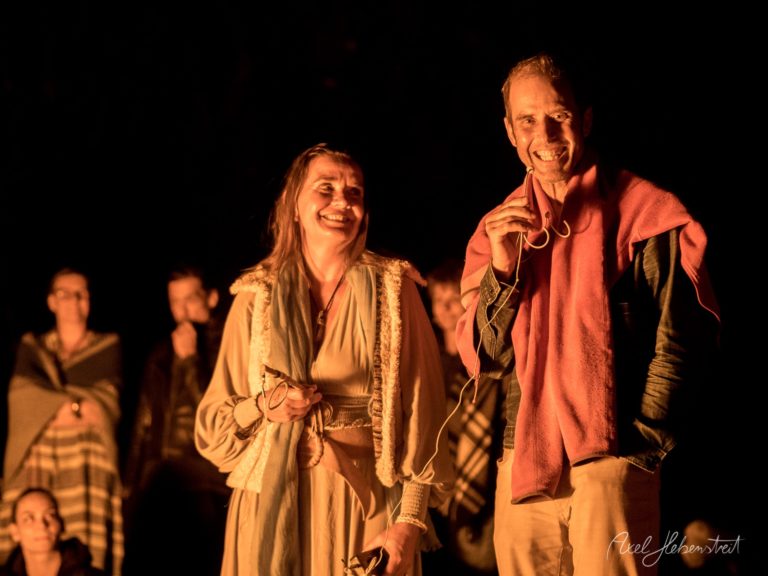 Zwei Performer strahlen sich auf einer Bühne an, umgeben von warmem Licht und Publikum im Hintergrund – © Axel Hebenstreit