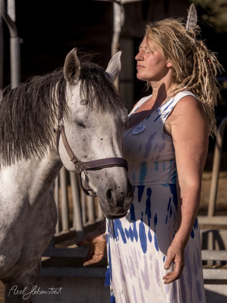 Frau mit Dreadlocks und Federn im Haar steht neben grauem Pferd im Stall und schaut verträumt zur Seite – © Axel Hebenstreit