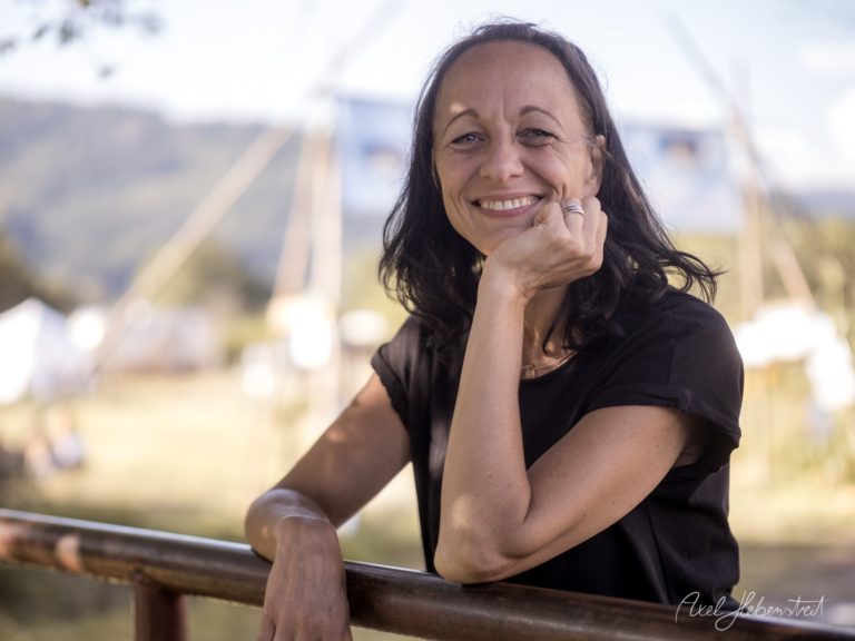 Lächelnde Frau lehnt an Geländer mit Flusslandschaft und Kirchtürmen im unscharfen Hintergrund – © Axel Hebenstreit