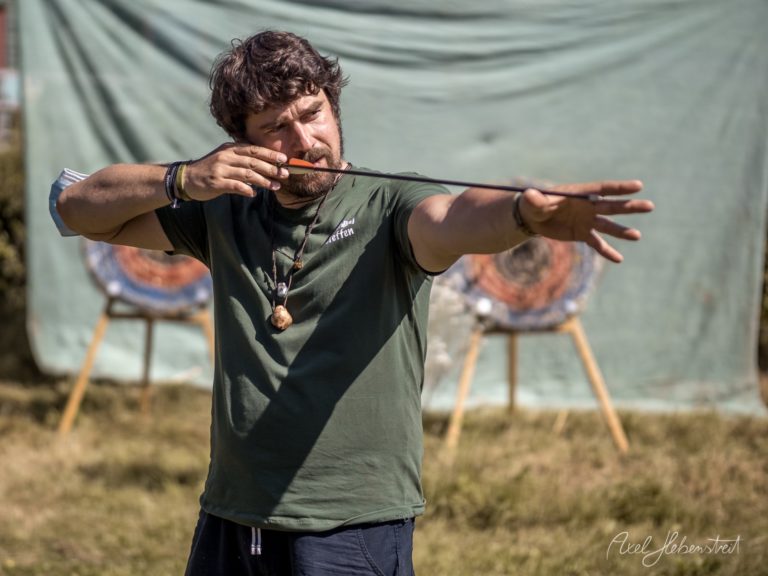 Mann demonstriert Bogenschießen auf Festival mit Zielen im Hintergrund – © Axel Hebenstreit