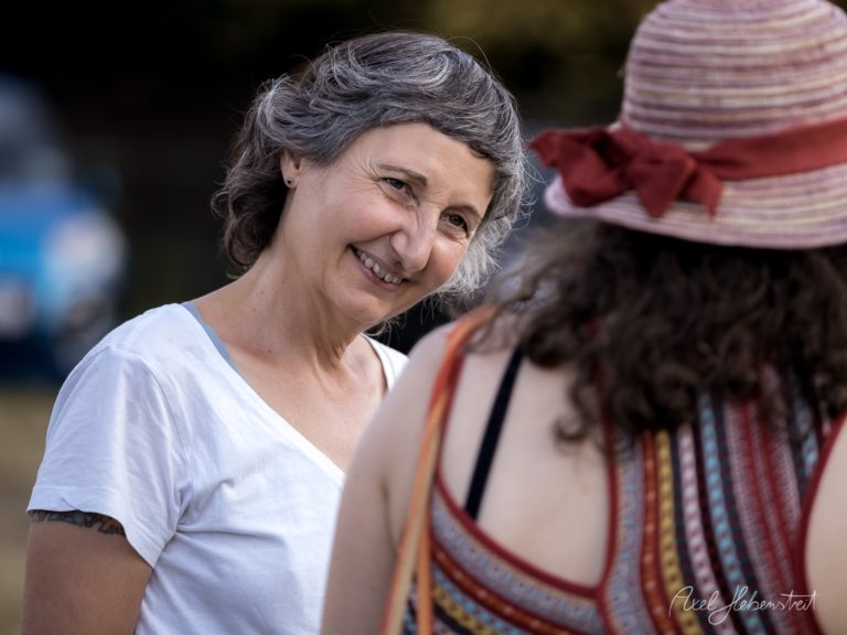 Lächelnde Frau mit grauem Haar im Gespräch mit traditionell gekleideter Person auf Festival – © Axel Hebenstreit