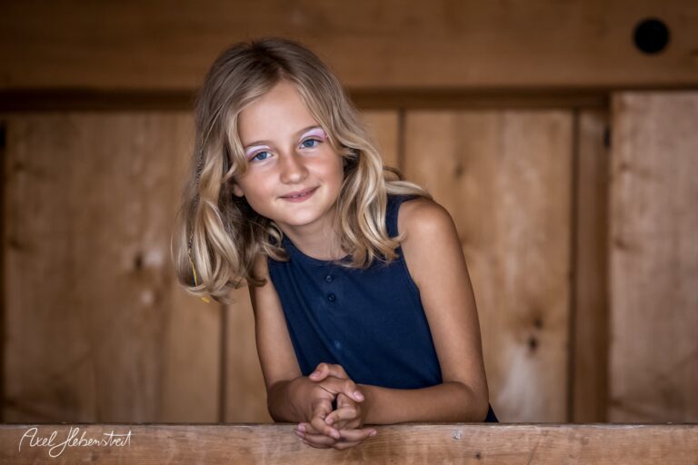 Ein lächelndes Mädchen mit blonden Locken steht an einem Holzgeländer in einem rustikalen Raum – © Axel Hebenstreit