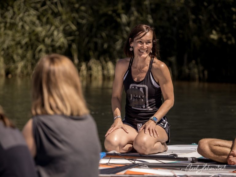 Eine lächelnde Frau sitzt auf einem Paddleboard am Wasser und spricht mit einer Gruppe von Zuhörern. – © Axel Hebenstreit