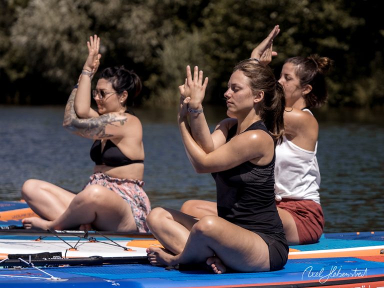 Drei Frauen üben Yoga auf Stand-Up-Paddle-Boards in einem ruhigen Gewässer – © Axel Hebenstreit