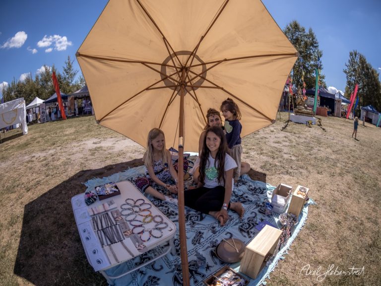 Eine Gruppe von Frauen sitzt unter einem Sonnenschirm an einem Stand auf einem Festival und präsentiert handgemachten Schmuck. – © Axel Hebenstreit