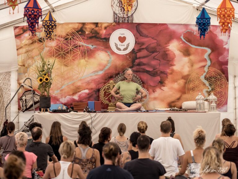 Eine entspannte Yoga-Session unter einem bunten Zelt mit Teilnehmern, die dem Lehrer auf der Bühne folgen – © Axel Hebenstreit