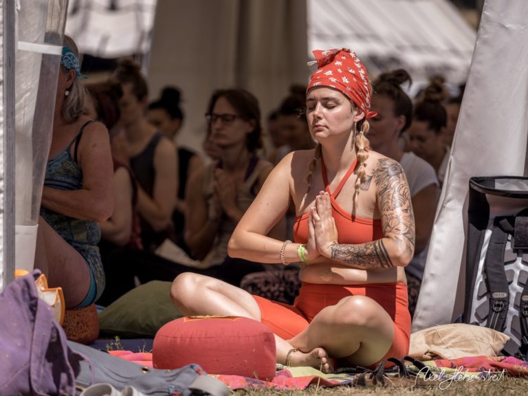 Eine yogainspirierte Frau mit Tattoos meditiert in roter Kleidung unter einem Zelt bei einem Festival. – © Axel Hebenstreit