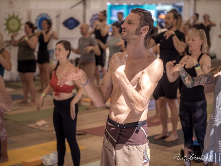 Ein Mann mit überlegener Mimik und einem entspannenden Ausdruck, umgeben von Teilnehmern in einer Yoga-Session – © Axel Hebenstreit