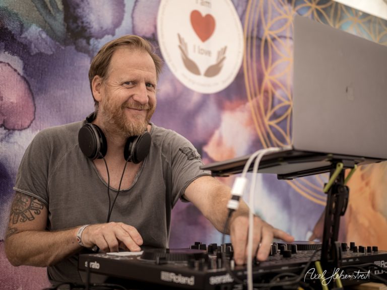 Ein DJ lächelt entspannt an seinem Pult in einer festlichen Umgebung mit bunten Hintergründen – © Axel Hebenstreit