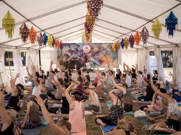 Yoga-Teilnehmerinnen und -Teilnehmer praktizieren im Zelt unter bunten Laternen bei einem Festival – © Axel Hebenstreit