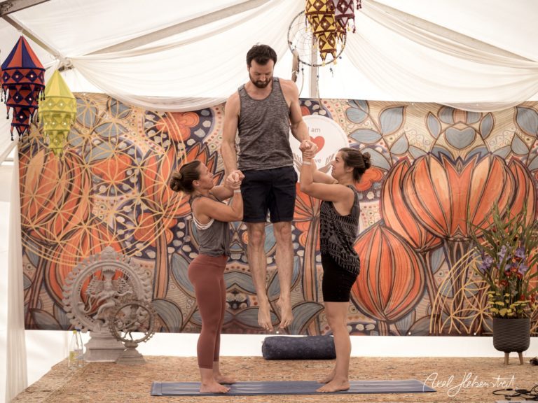 Zwei Frauen unterstützen einen Mann beim Yoga, während er in der Luft schwebt, vor einer bunten Wand im Festivalambiente. – © Axel Hebenstreit