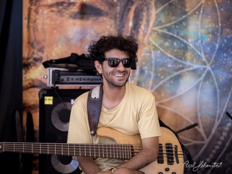Lächelnder Musiker mit einem E-Bass sitzt entspannt vor einer bunten Festivalkulisse – © Axel Hebenstreit