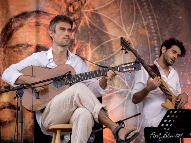Zwei Musiker, einer mit Gitarre und der andere am Bass, spielen konzentriert auf einer Festivalbühne – © Axel Hebenstreit
