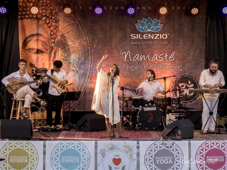 Eine Sängerin performt energetisch auf einer Bühne, umgeben von Musikern in entspannter Festivalstimmung – © Axel Hebenstreit